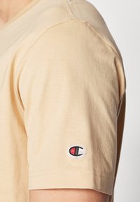 Beiges T-Shirt mit kurzen Ärmeln aus Baumwolle, mit Rundhalsausschnitt und kleinem gesticktem Logo in Rot, Schwarz und Weiß am Ärmel.
