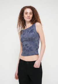 Γυναίκα με σγουρά κόκκινα μαλλιά που φοράει μπλε μπλουζάκι με λουλούδια χωρίς μανίκια και μαύρο παντελόνι, με το ένα χέρι στην τσέπη, στέκεται μπροστά σε απλό φόντο.