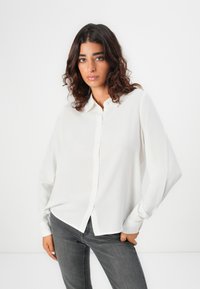 Camicia bianca con bottoni, maniche lunghe, realizzata in tessuto leggero, abbinata a jeans grigi. Caratterizzata da un colletto classico e una vestibilità rilassata.