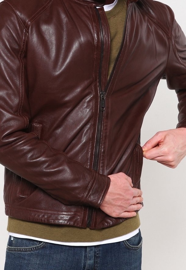 Braune Lederjacke mit Reißverschluss, seitlichen Taschen und gerippten Bündchen. Sie hat eine glatte Textur und eine taillierte Passform. Getragen über einem grünen Oberteil.