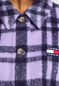 Fialová a černá kostkovaná flanelová košile s límcem, obsahující nášivku s logem a zapínání na značkový knoflík. Měkká textura a upravený střih.