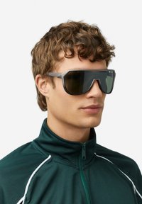 Športové slnečné okuliare so sivým plastovým rámom, tmavými tónovanými sklami a uhlovým dizajnom. Nosené modelom v zelenej bunde na zips.