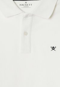Witte polo met een gestructureerde stof. Beschikt over een traditionele kraag, twee knopen en een klein zwart logo met gekruiste voorwerpen op de borst.