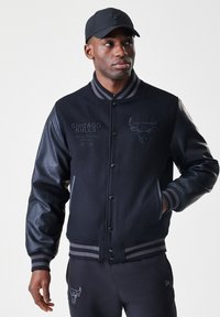 Homem de casaco varsity preto do Chicago Bulls e boné, olhando para o lado com uma mão no bolso contra um fundo simples.