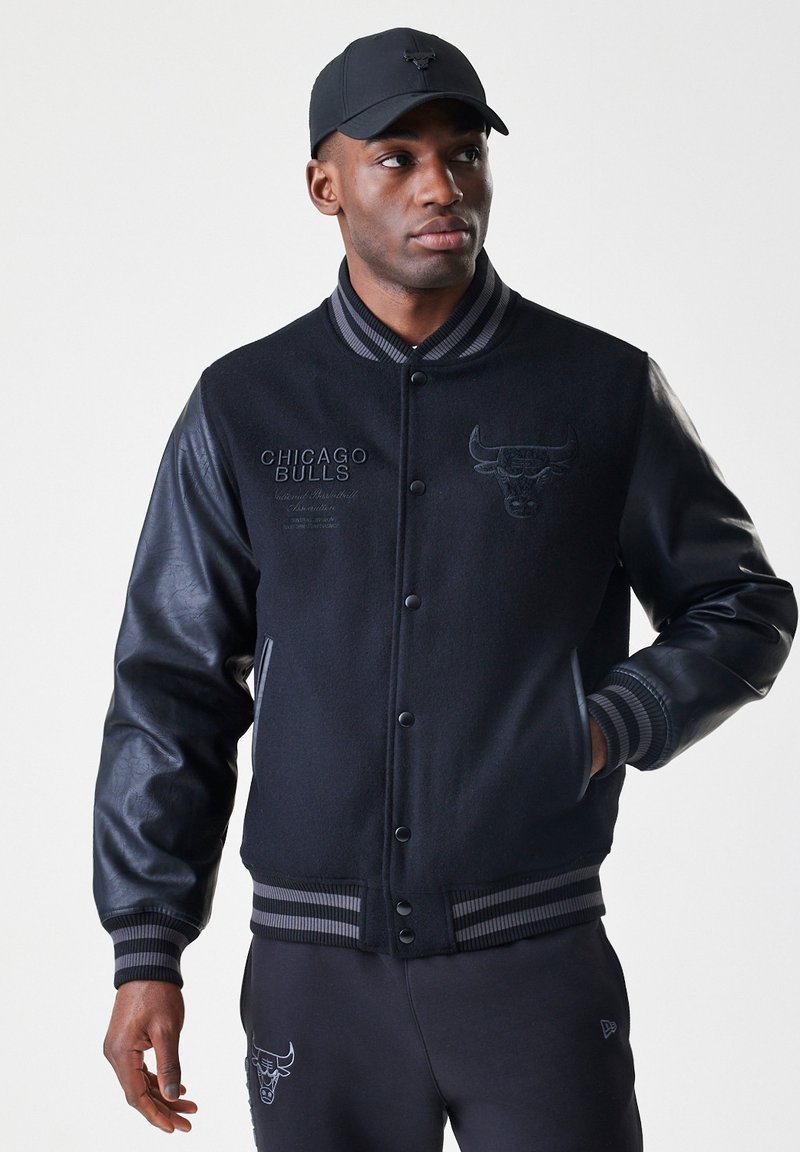 Homem de casaco varsity preto do Chicago Bulls e boné, olhando para o lado com uma mão no bolso contra um fundo simples.