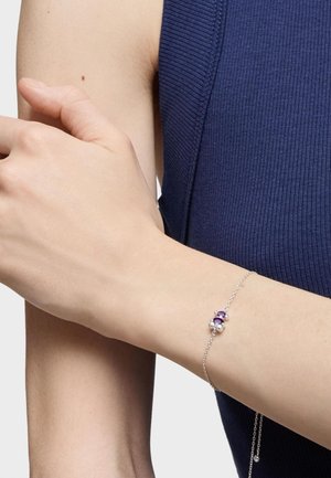 Bracciale d'argento con gemme viola e bianche, dotato di una delicata catena e un semplice design ispirato ai fiori. Indossato su un polso nudo.