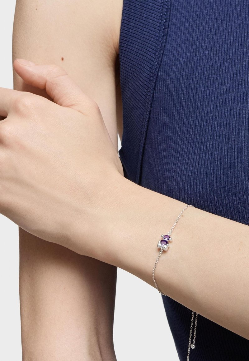 Pulsera de plata con piedras preciosas moradas y blancas, con una delicada cadena y un sencillo diseño inspirado en flores. Lleva en una muñeca desnuda.