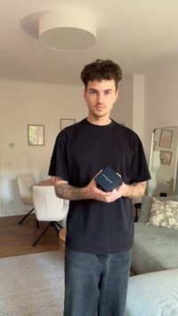 Cutie neagră închisă, ținută în mâini, cu un finisaj mat și litere albe, așezată într-o cameră luminată, cu mobilă și decor în culori neutre.