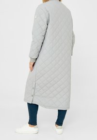 Donna che indossa un lungo cappotto trapuntato grigio chiaro con bottoni a pressione sul lato, jeans blu scuro e sneakers bianche, in piedi su uno sfondo bianco.