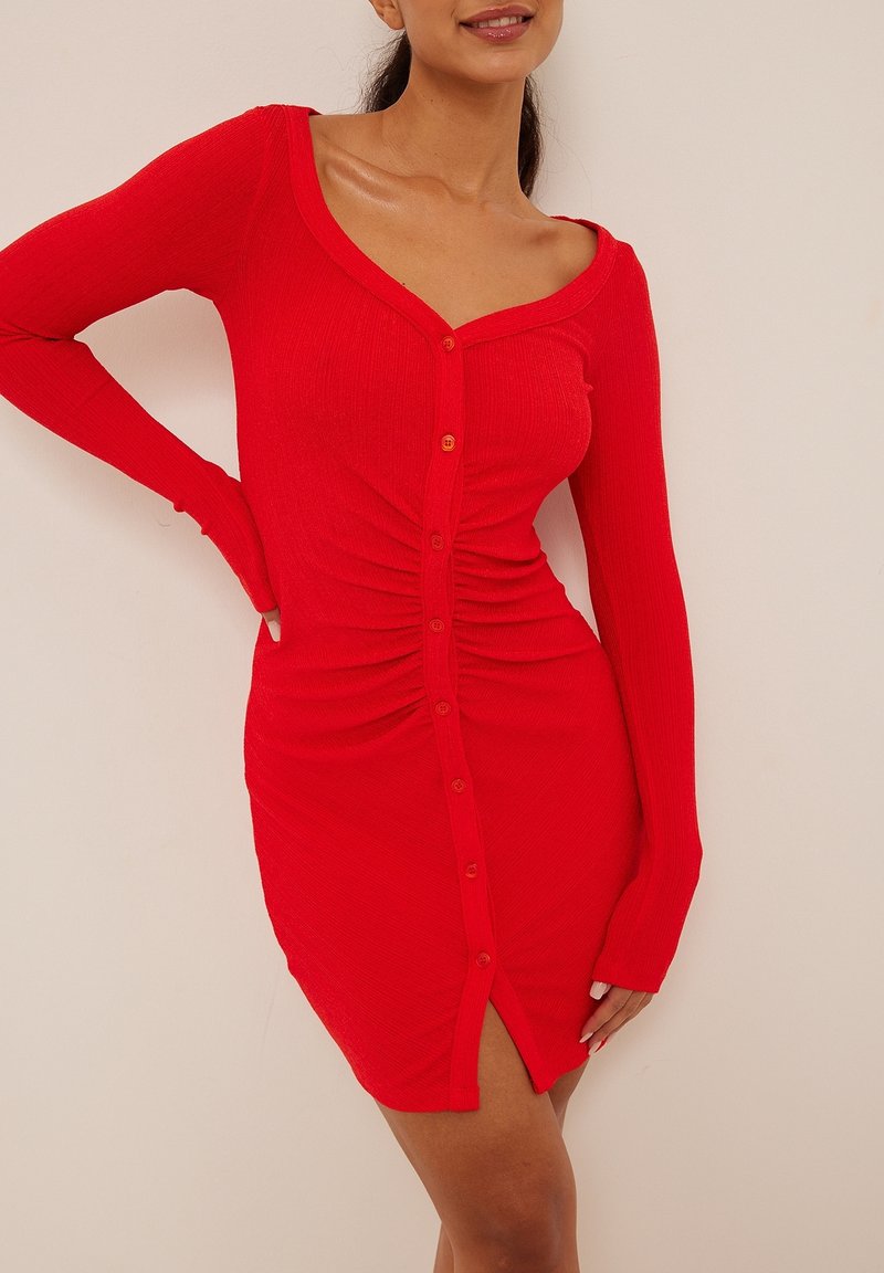 Robe rouge côtelée à manches longues, avec un décolleté en V et une fermeture boutonnée au centre, des détails froncés sur les côtés, et une silhouette ajustée.