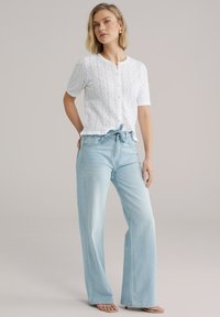 Top bianco a maniche corte con texture in pizzo e chiusura con bottoni; abbinato a jeans larghi azzurri con vita a coulisse.