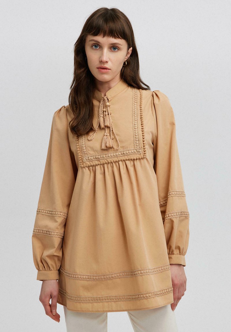 Beige tunika med høj halsudskæring, samlet detaljering og pufærmer. Har dekorativ syning og frynser foran.