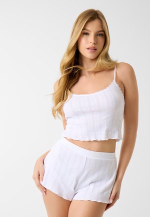 Jonge vrouw met lang blond haar, gekleed in een wit, gestructureerd mouwloos crop top en bijpassende shorts met hoge taille, staand tegen een effen achtergrond.