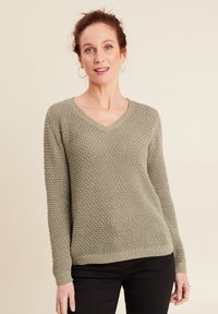 V-hals trui in licht olijfgroen met een gebreide structuur, lange mouwen en een geribbelde zoom. Zwarte broek maakt de outfit compleet.