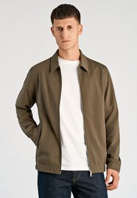 Oliwkowo-zielona kurtka na zamek z kołnierzem, lekka tkanina, boczne kieszenie, w zestawieniu z białym t-shirtem i ciemnymi dżinsami. Gładka struktura.