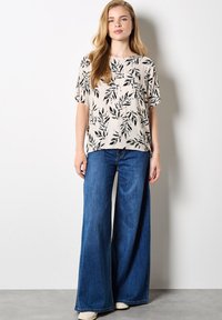 Lettet beige bluse med sort bladmønster, korte ærmer, rund halsudskæring, parret med vidde denim jeans i blå. Enkel baggrund.