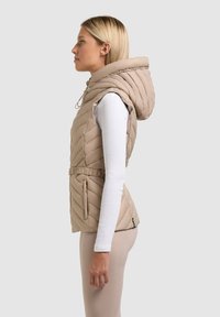 Vrouw in beige gewatteerd mouwloos gilet met capuchon over wit longsleeve, staand in profiel tegen een effen achtergrond.