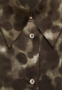 Brown short-sleeve shirt with a collar, featuring a spotted abstract pattern in darker and lighter shades. Single mother-of-pearl button detail.