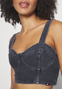 Sötét szürke farmer crop top, struktúrált bögre fazonnal, széles pántokkal és kontrasztos varrással. Kisebb logó címkével rendelkezik piros, fehér és kék színekben.