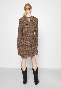 Kvinna med blont hästsvans som bär en långärmad klänning i brunt och svart mönster, svarta cowboystövlar, vänd bort mot en enkel bakgrund.