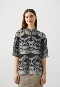 Blusa nera con motivi geometrici argentati, maniche a tre quarti e scollatura a goccia. Il dettaglio dell'orlo scallop aggiunge texture.