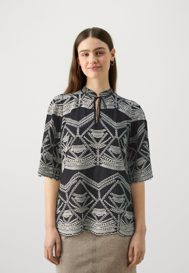 Blusa preta com padrões geométricos prateados, mangas três quartos e decote em forma de chave. Detalhe de bainha recortada adiciona textura.