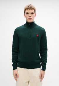 Grüner Strickpullover mit hohem Kragen und halbem Reißverschluss. Verfügt über ein kleines rotes Logoschild auf der Brust. Kombiniert mit hellen Hosen.