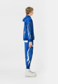 Sudadera azul con capucha y pantalones de chándal que presentan diseños detallados de alas. El material es suave, con una textura lisa. Bolsillos acentuados en blanco. Botas blancas.