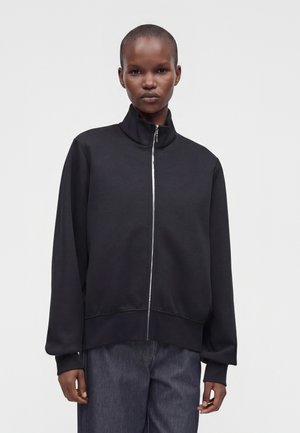 Personne aux cheveux courts portant une veste noire zippée et un pantalon foncé, debout devant un fond clair uni.