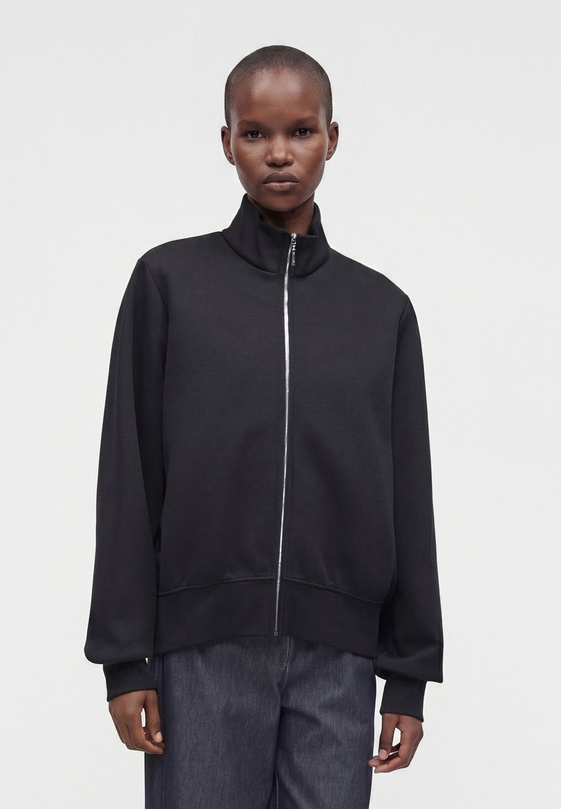 Personne aux cheveux courts portant une veste noire zippée et un pantalon foncé, debout devant un fond clair uni.