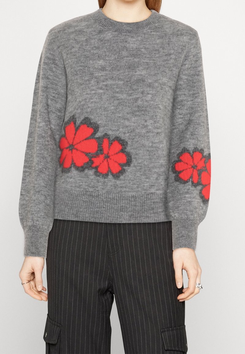 Pull gris en maille avec des motifs floraux rouges sur le devant et les manches, porté avec un pantalon noir à fines rayures et poches latérales.