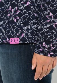 Närbild av hand bredvid mörkjeans och svart långärmad tröja med vit och rosa geometrisk blommönster samt rosa logotypmärkning.