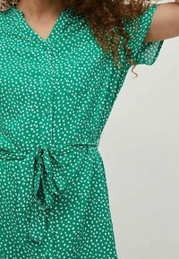 Grön klänning med v-ringning, korta ärmar och resårlinning i midjan. Tyget har ett vitt abstrakt mönster och en lätt textur.