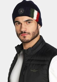 Beanie lavorato a maglia blu navy con un logo circolare e una fascia tricolore verde, bianca e rossa. Indossato con un gilet nero imbottito sopra una camicia bianca.