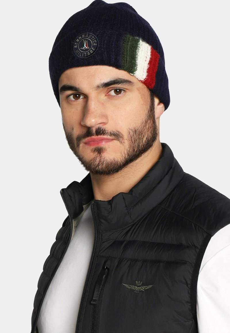 Beanie lavorato a maglia blu navy con un logo circolare e una fascia tricolore verde, bianca e rossa. Indossato con un gilet nero imbottito sopra una camicia bianca.
