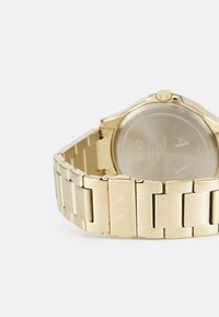 Reloj de metal dorado con pulsera de eslabones y caja redonda. La parte trasera es transparente, mostrando la mecánica interna del reloj. Presenta una corona.