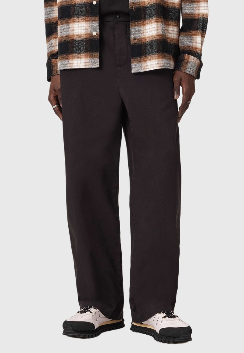Pantaloni neri a gamba ampia in cotone, con una trama liscia e un taglio classico. Indossati con una camicia a quadri con bottoni e sneakers.