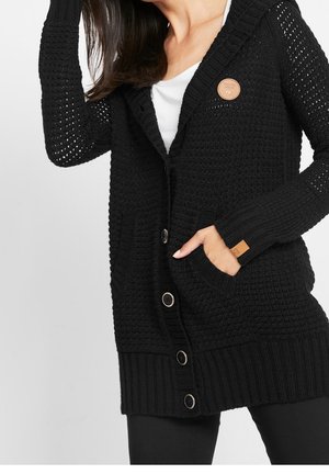 Femme portant un cardigan en maille noire avec de gros boutons, une main dans la poche, par-dessus une chemise blanche.