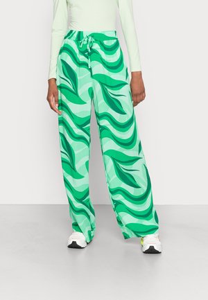 Person wearing wide-leg pants with green abstract leaf patterns and white sneakers, standing against a plain background.
