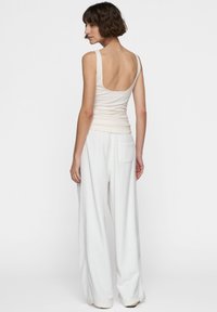 Mujer con cabello corto castaño, vistiendo una blusa sin mangas beige con la parte de atrás baja y pantalones blancos holgados, de pie contra un fondo blanco liso.