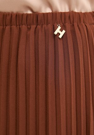 Gonna plissettata marrone con elastico in vita e piccolo ciondolo a forma di lettera "H" dorata attaccato vicino alla parte superiore centrale.