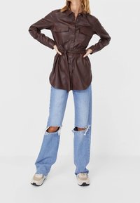 Brązowa sukienka koszulowa z eko skóry z zapięciem na guziki, dwoma kieszeniami na piersiach, paskiem w talii, zestawiona z przetartymi jasnoniebieskimi dżinsami i beżowymi trampkami.