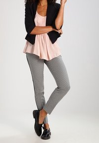 Zwarte blazer over een lichtroze geplooide top, gecombineerd met strakke zwart-witte patroonbroeken en zwarte schoenen.