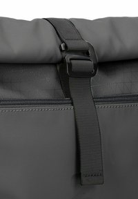 Bolsa impermeable gris con cierre enrollable, hebilla de plástico negra y bolsillo con cremallera que presenta una textura de patrón geométrico.