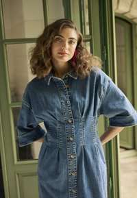 Denimkleit, millel on nupuväli, kraega kaelus ja poolpikad varrukad. Kangas on keskmise pesu efektiga, pehme tekstuuriga ja volditud detailidega.