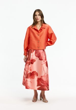 Frau trägt eine glänzende orangene Jacke mit Knöpfen und einen rosa geblümten Midirock, steht mit einer Hand in der Hüfte und trägt beigefarbene offene Sandalen.