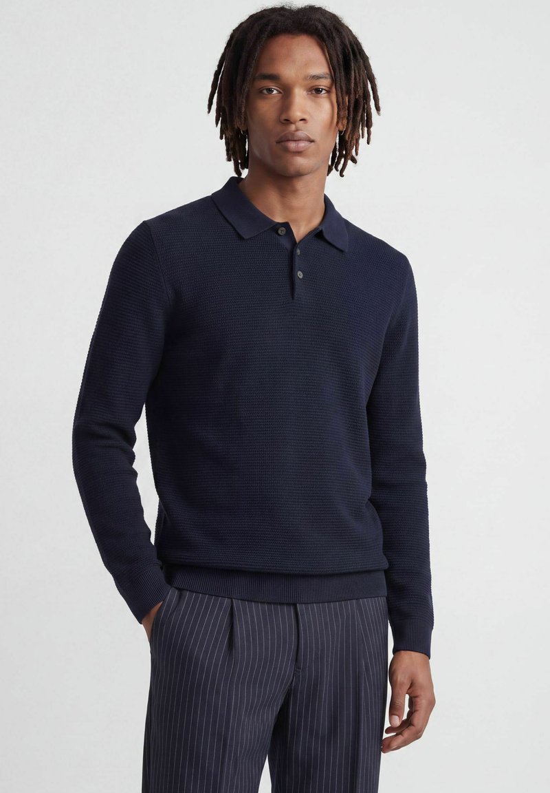 Jeune homme avec des dreadlocks de longueur moyenne portant un polo texturé bleu marine à manches longues et un pantalon sombre à fines rayures, debout devant un fond uni.