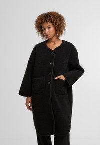 Cappotto nero, oversize, realizzato in un tessuto testurizzato e soffice, con scollo a V, chiusura con bottoni e due tasche frontali. Presenta maniche lunghe.