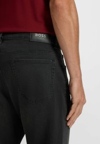 Pantaloni in denim nero con una texture liscia, dotati di un patch con il logo BOSS sulla vita e una singola tasca posteriore.
