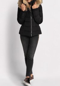 Doudoune noire avec capuche en fausse fourrure, fermeture éclair et design matelassé, associée à un jean foncé et des baskets marron.